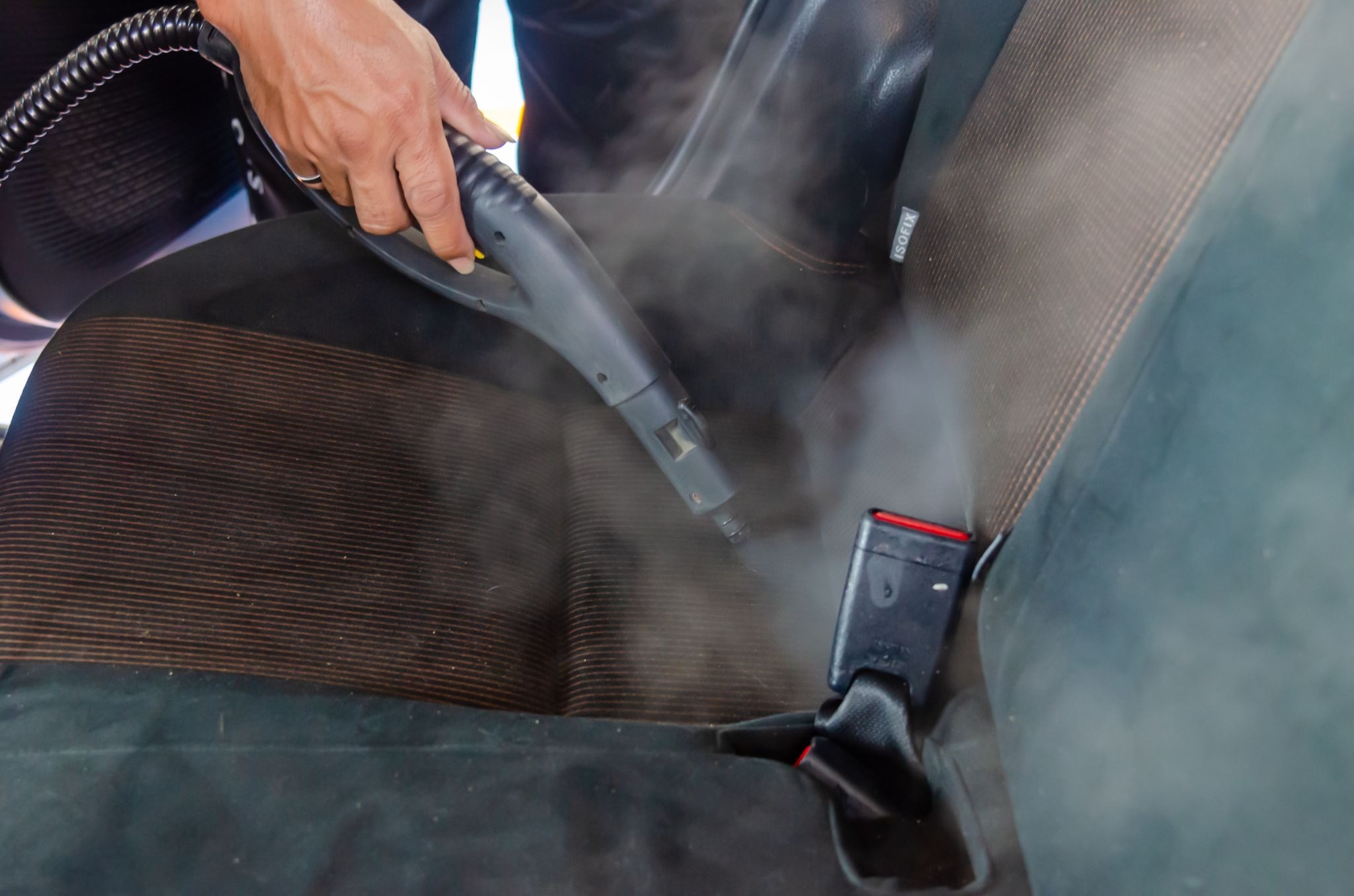 A person uses a steam cleaner to sanitize a car seat, with visible steam clearly emanating from the nozzle