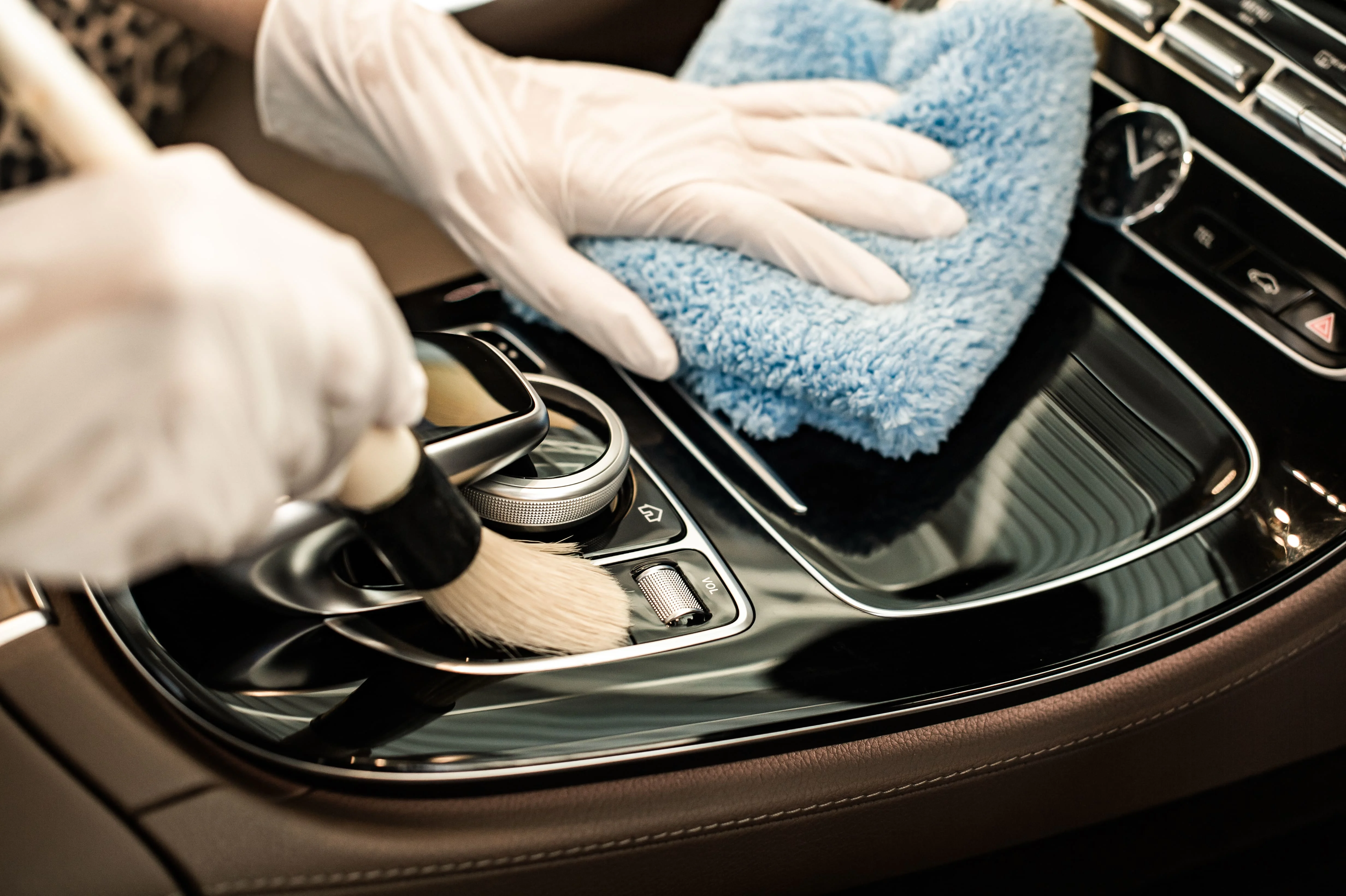 A person wearing white gloves cleans a car’s gear console using a blue cloth and a brush