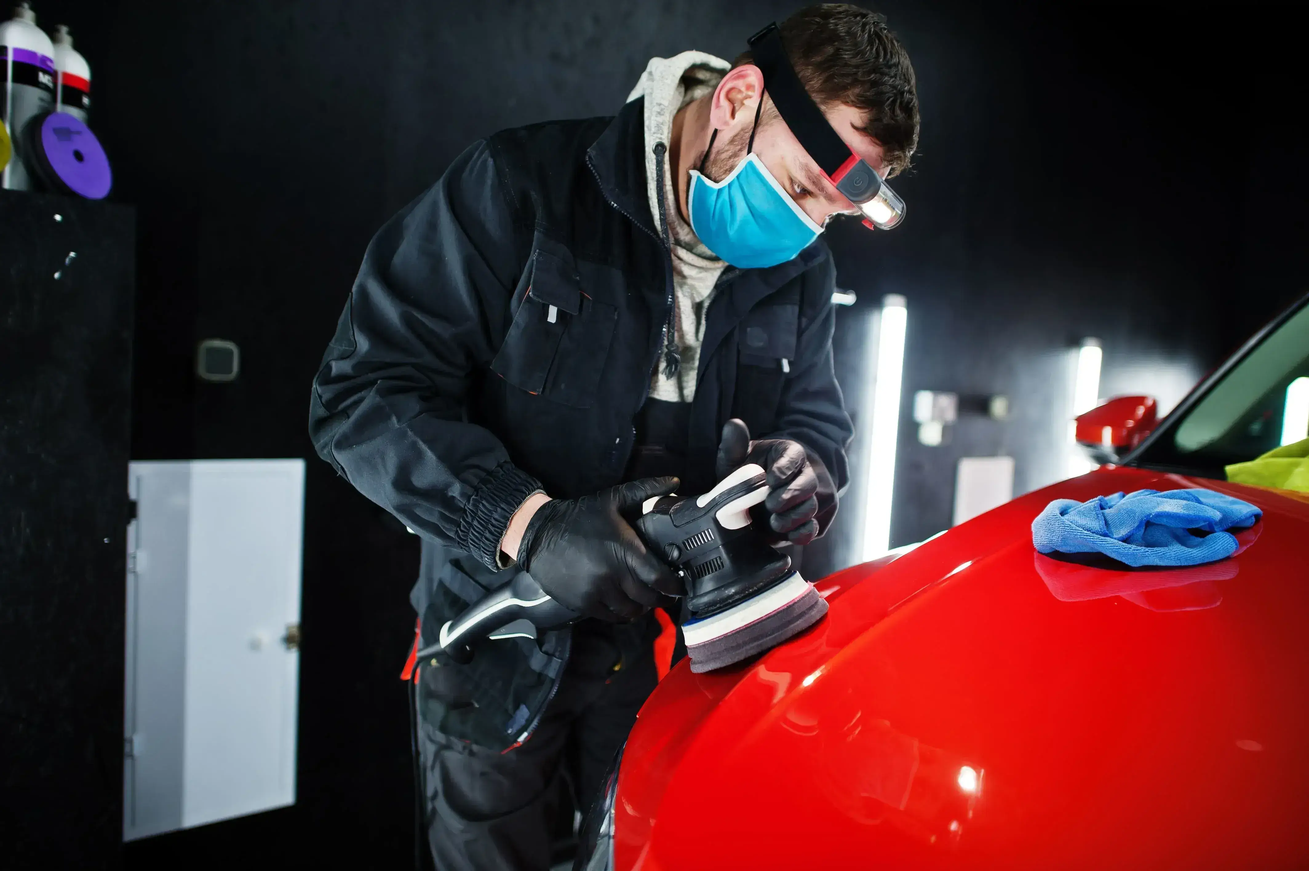 A person wearing a mask and a headlamp polishes the hood of a red car